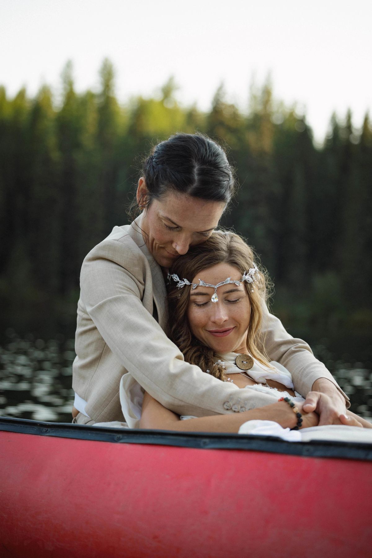 Two brides holding each other in a red canoe on a still forest lake