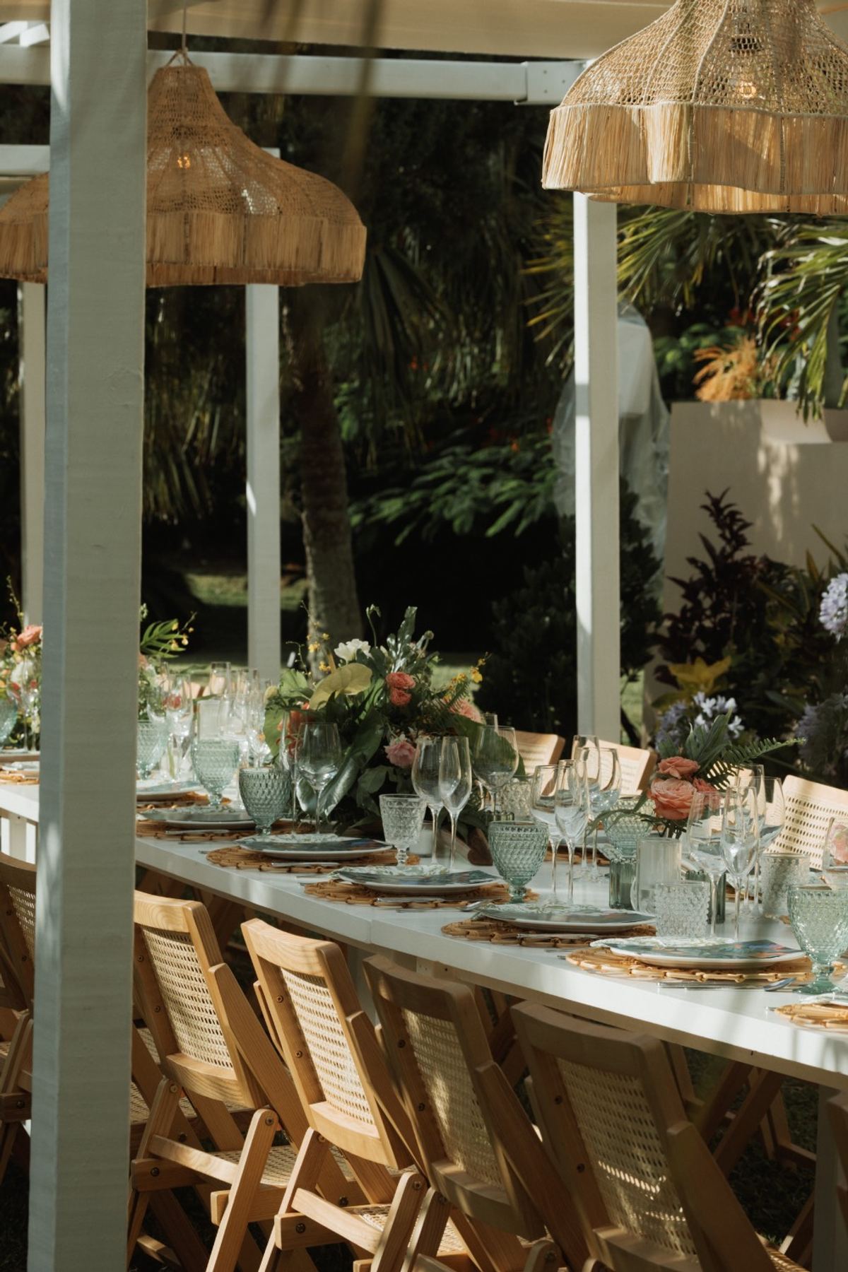 Long reception table laid with green glassware and blooms under a thatched pergola