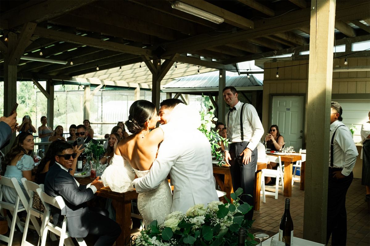Couple kissing at their table under a timber pavilion as guests look on