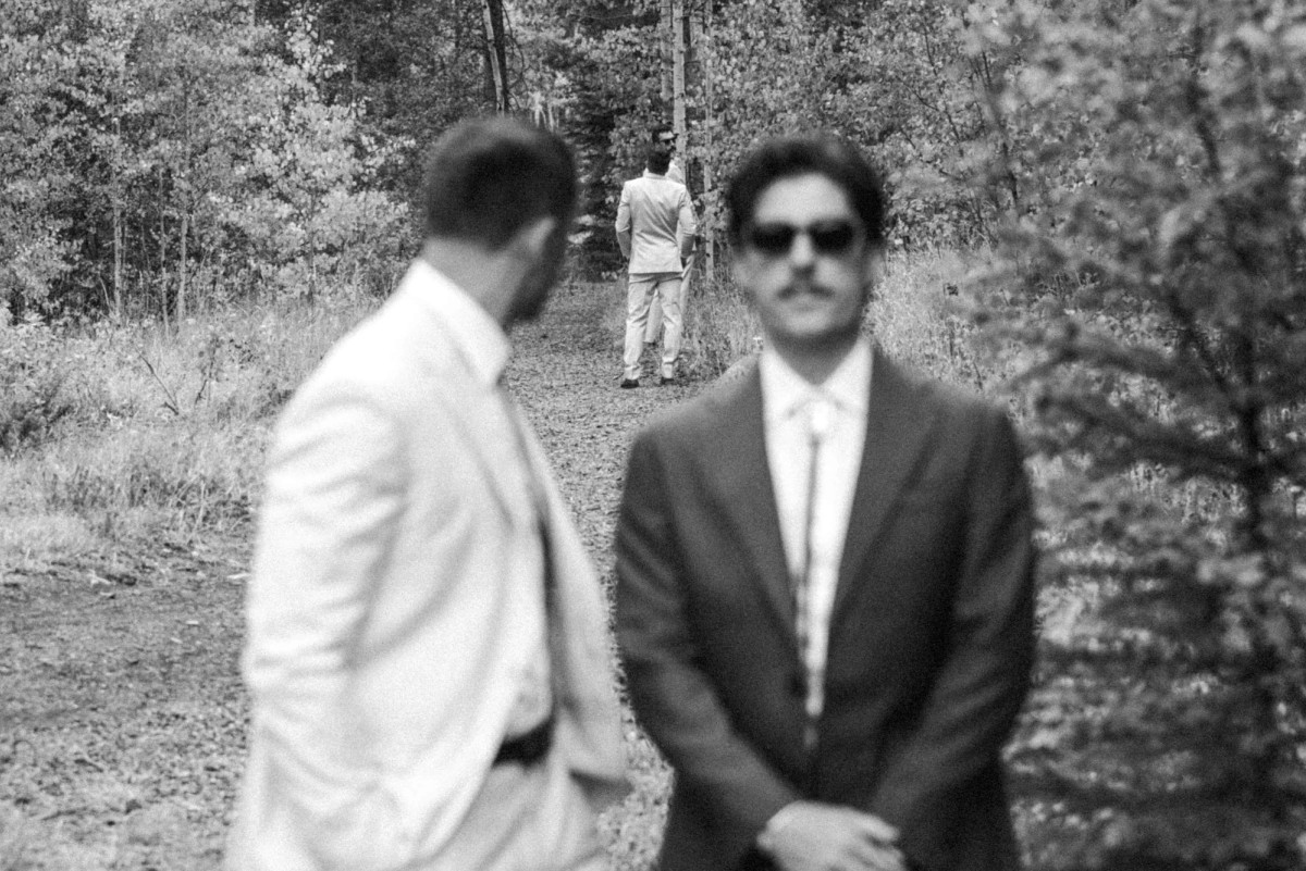 Owen waits in sunglasses as a groomsman looks back down the trail at a mountain ranch among the aspens, black and white