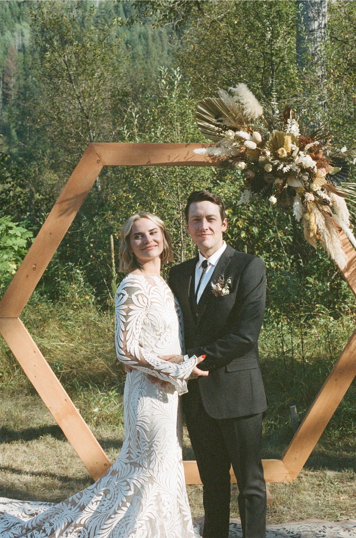 Bride and groom beneath a hexagonal wooden arch dressed with pampas grass