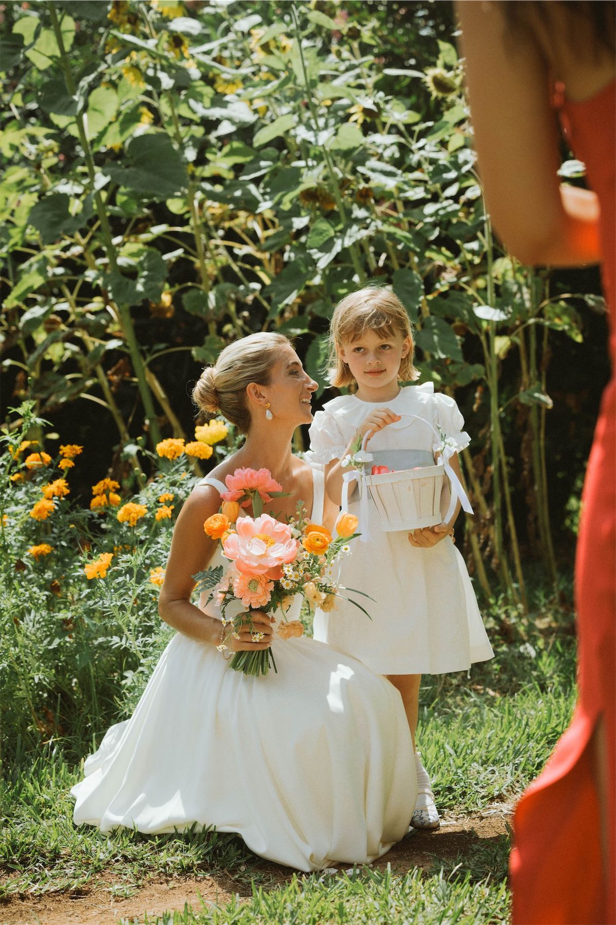 Bride kneeling beside the flower girl with a marigold bouquet in hand