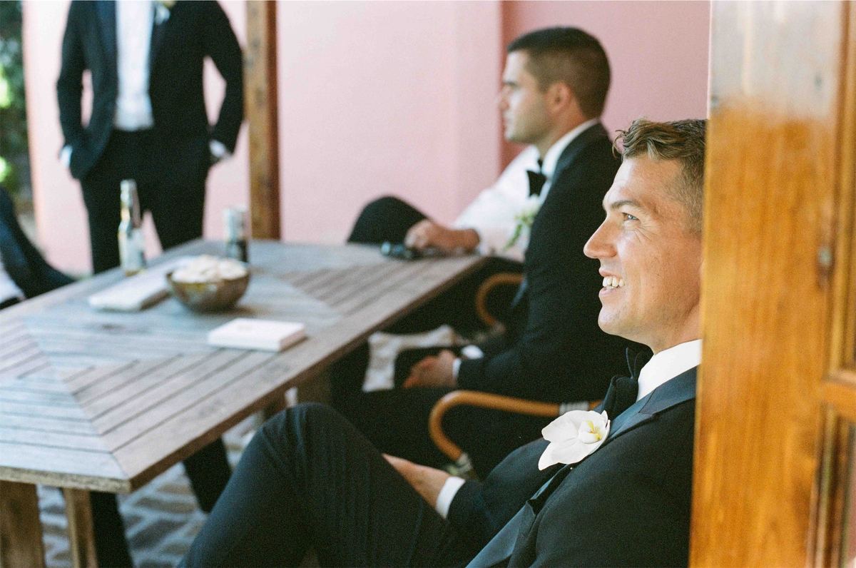 Groom relaxing at a table with his groomsmen against a coral pink wall