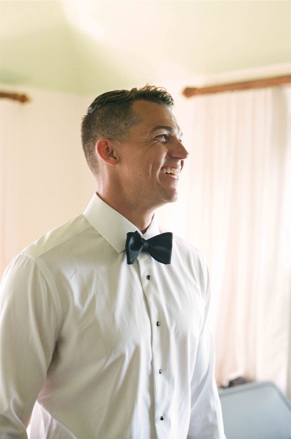 Groom laughing in shirt and bow tie while getting ready in a sunlit room