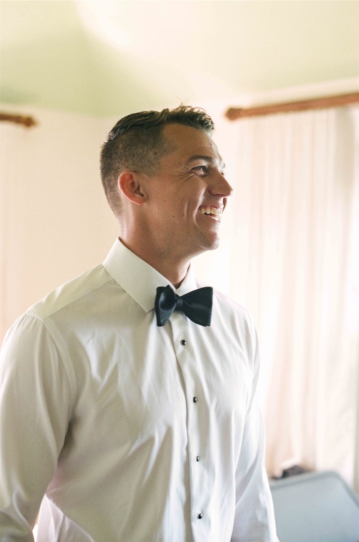 Groom laughing in shirt and bow tie while getting ready in a sunlit room