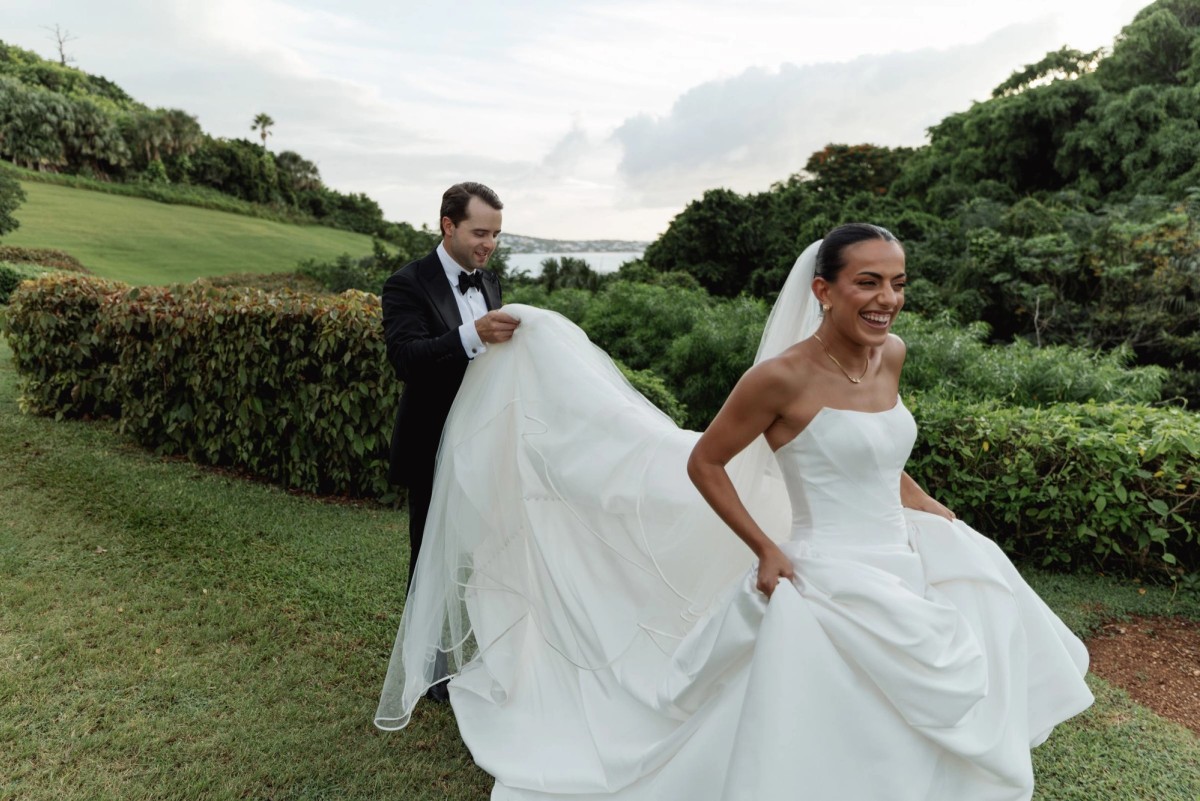 Casey carries Juliana's long train across a green hillside above the sea at Rosewood Bermuda