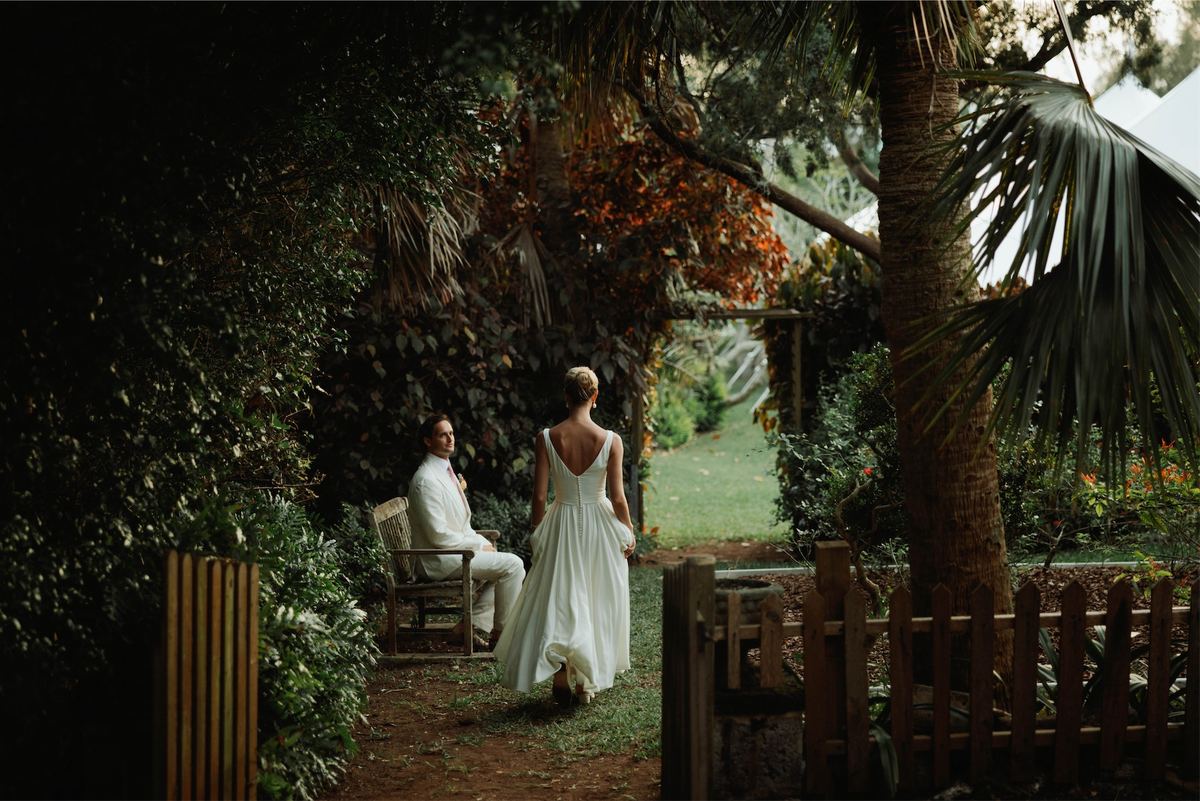 Bride in a low-back gown walking through a garden gate toward the groom