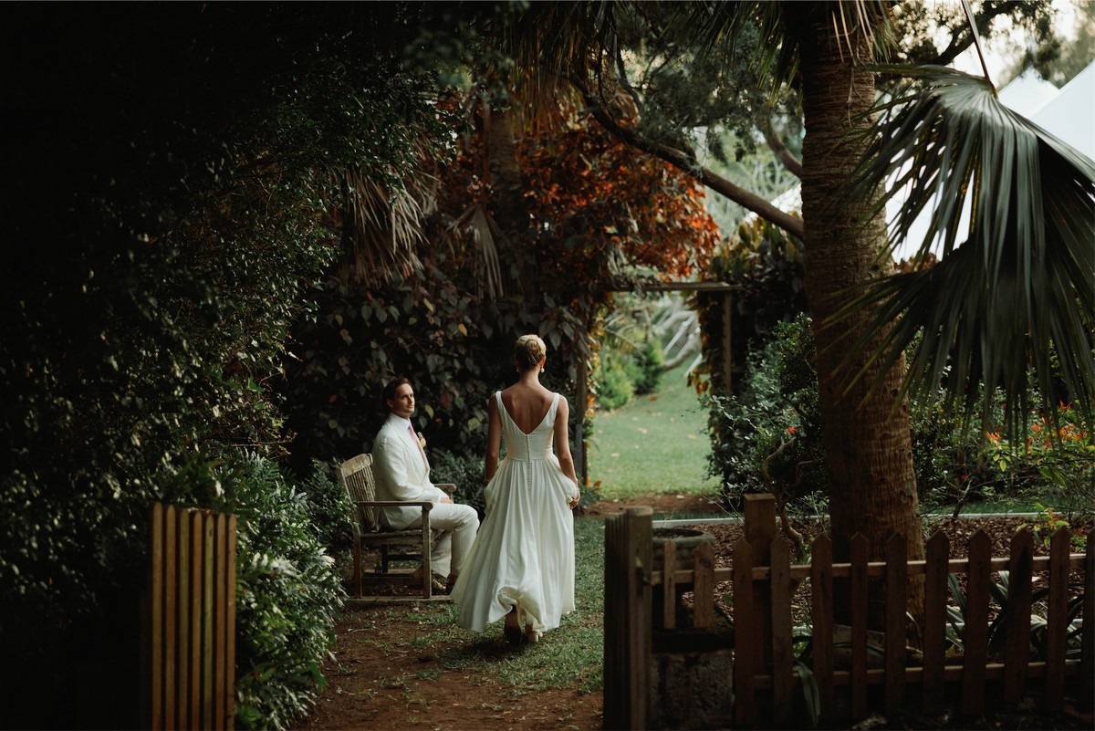 Bride in a low-back gown walking through a garden gate toward the groom