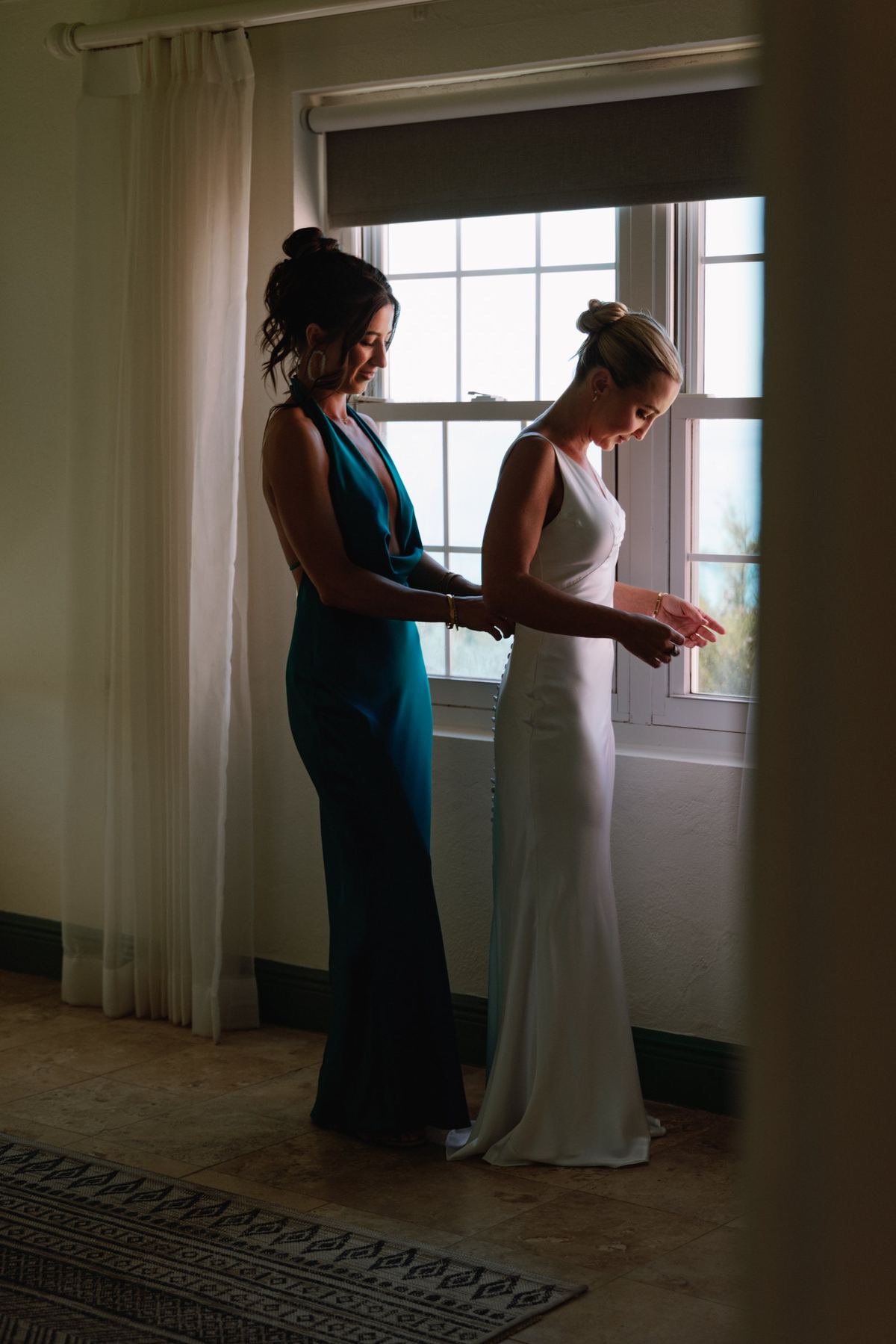 Bride being helped into her gown beside a bright window before her Bermuda wedding