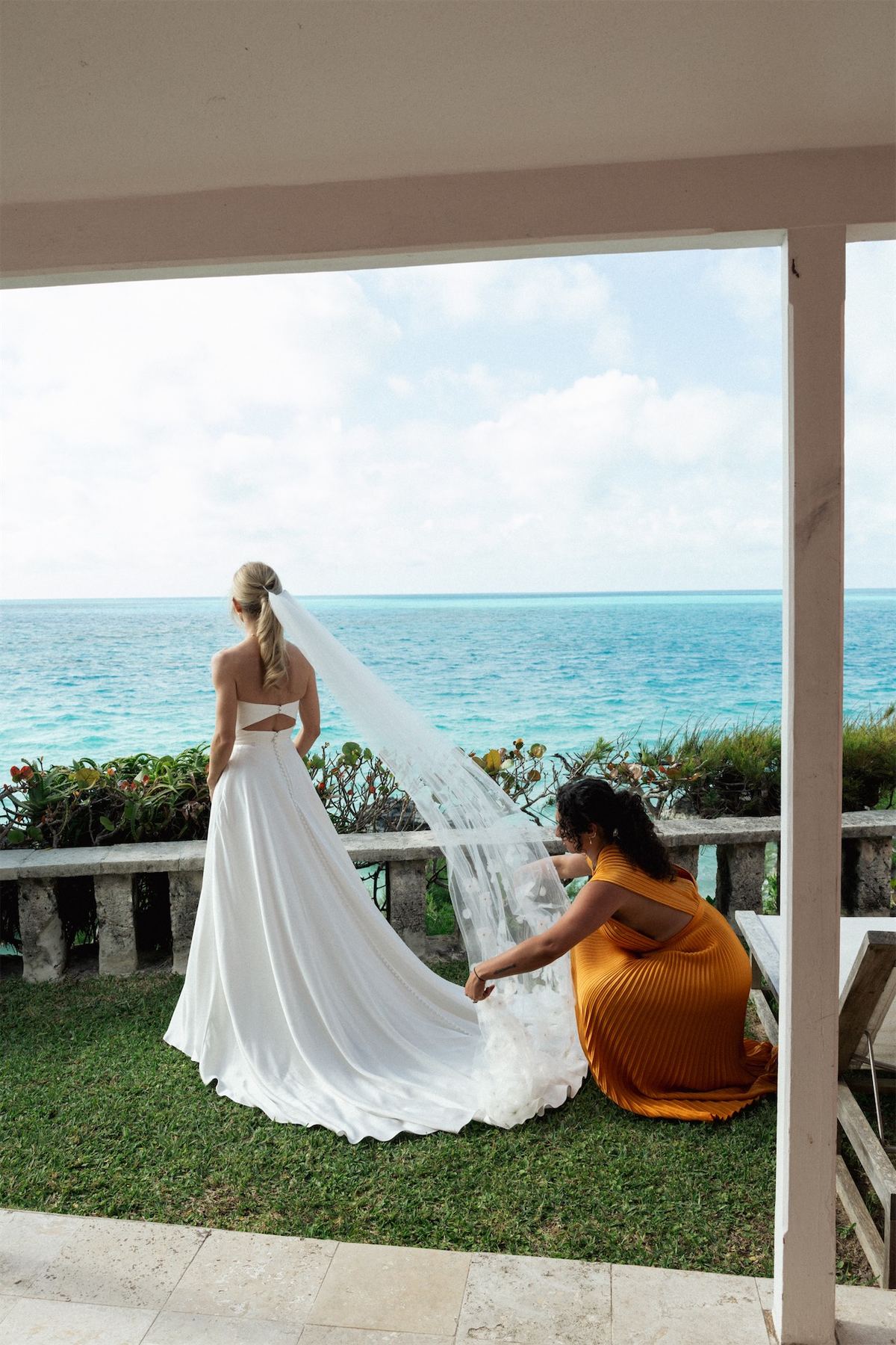 Bride's veil and train being arranged on a terrace above turquoise water