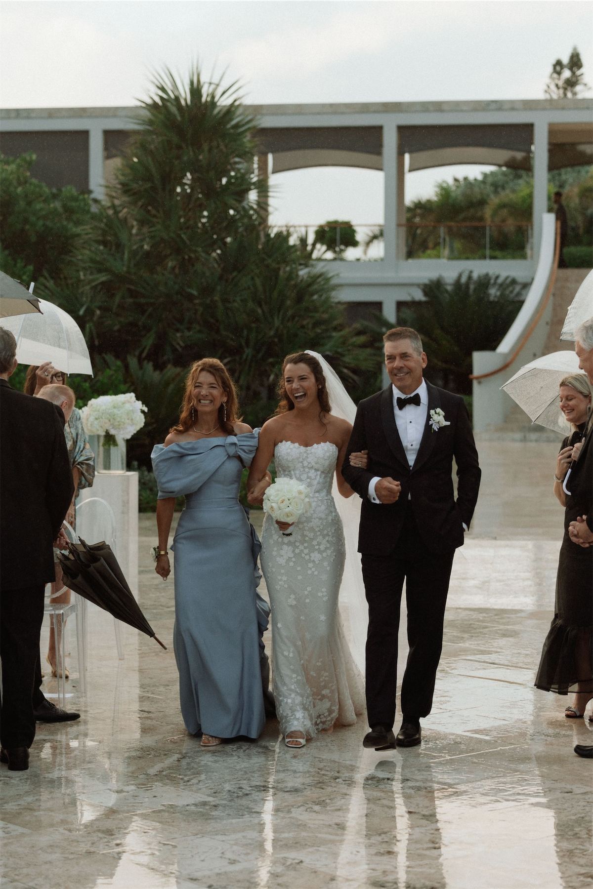 Bride arriving arm in arm with her parents past guests holding umbrellas