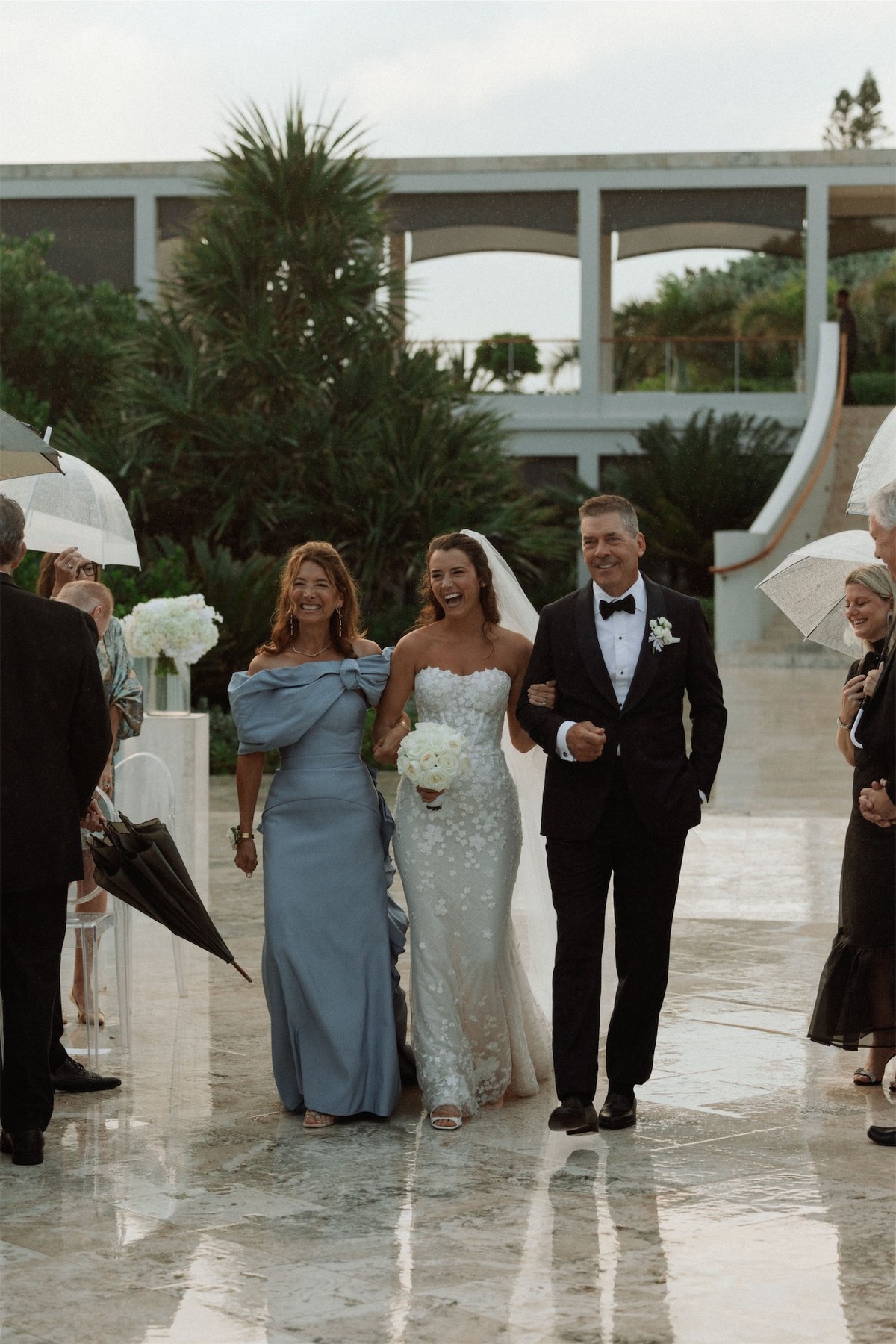 Bride arriving arm in arm with her parents past guests holding umbrellas