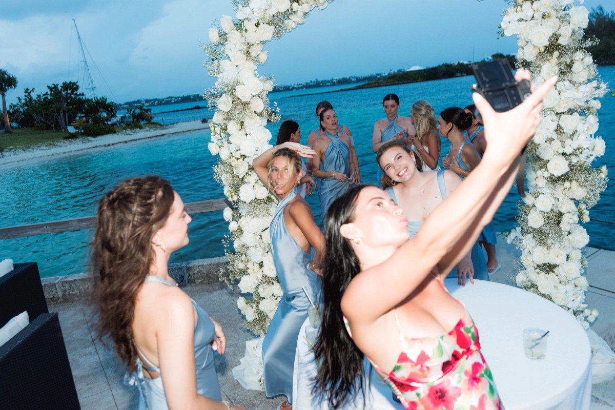A guest takes a selfie with the bridesmaids under the floral arch on Hawkins Island, Bermuda