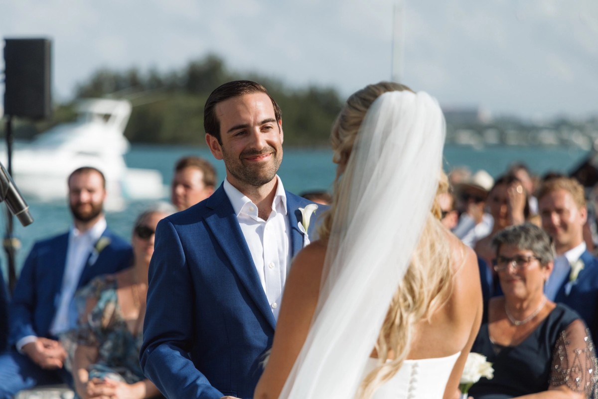 Cam smiles at Paige during the vows with guests and boats behind on Hawkins Island, Bermuda