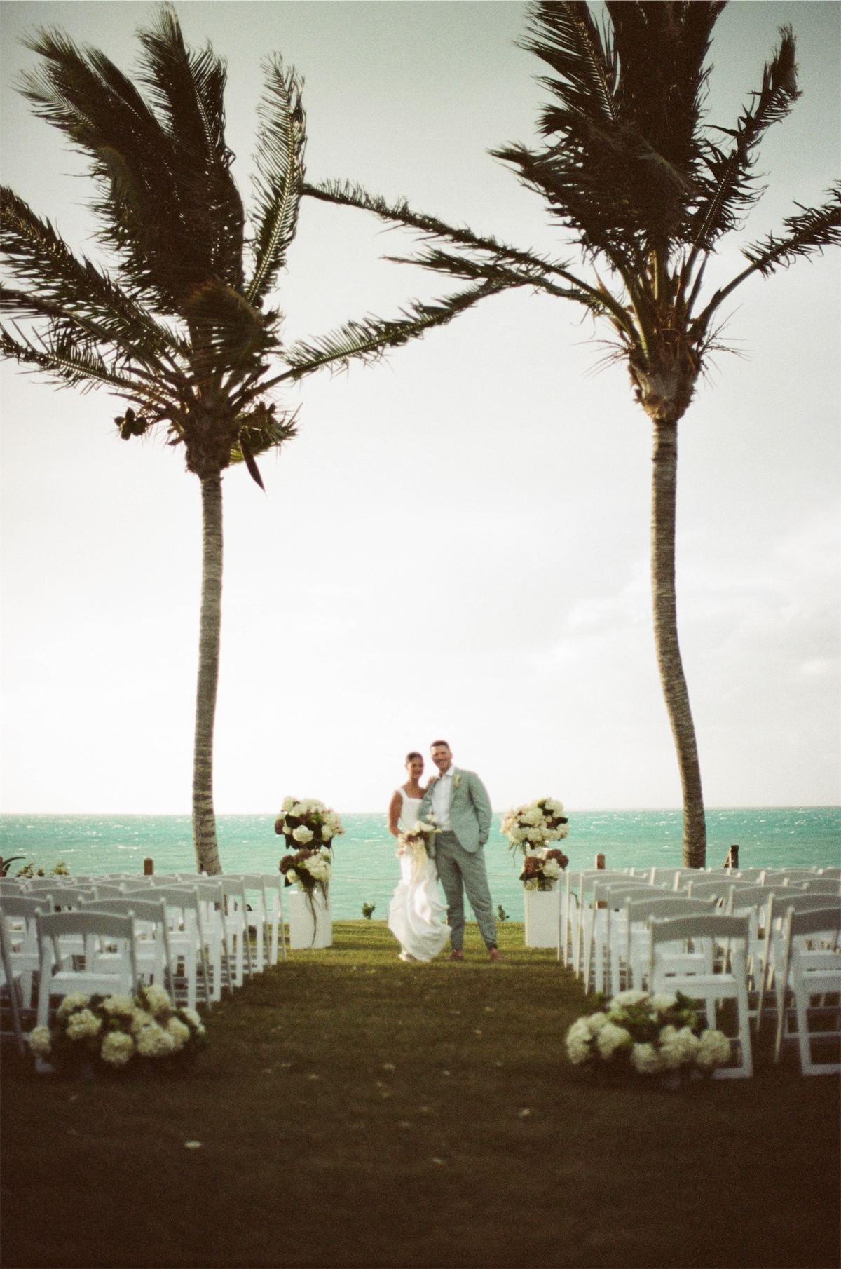 Ceremony set on a lawn between two leaning palms with the ocean beyond