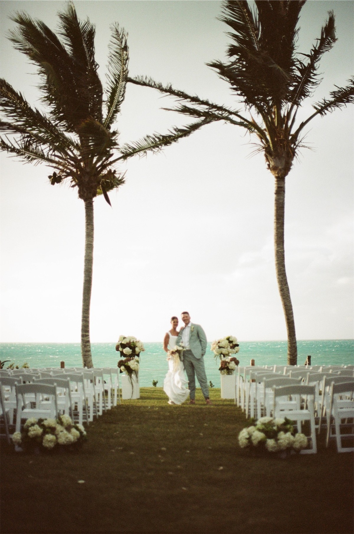 Ceremony set on a lawn between two leaning palms with the ocean beyond