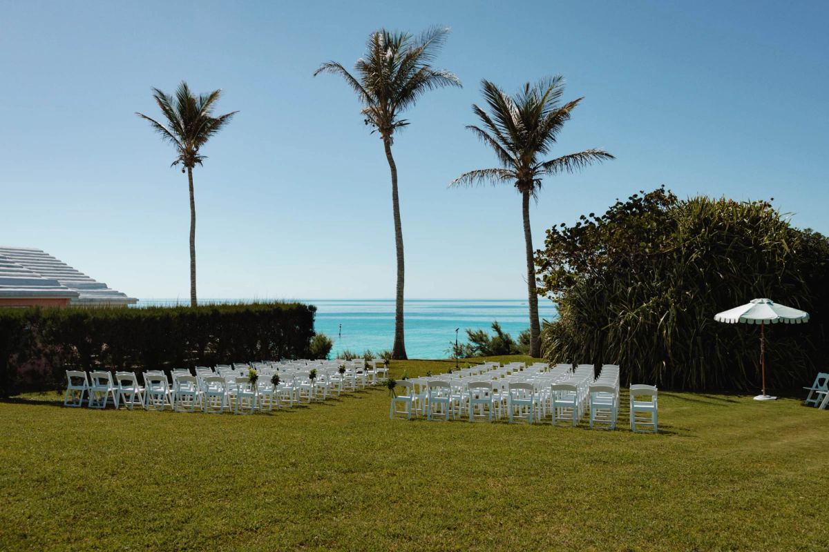Empty ceremony chairs set on the lawn between palm trees above the Bermuda ocean