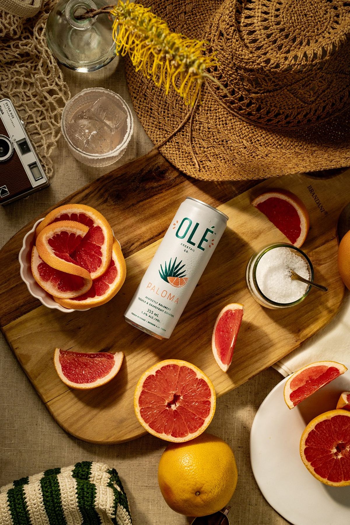 Overhead flat lay of a canned margarita on a wood board with sliced grapefruit and a straw hat