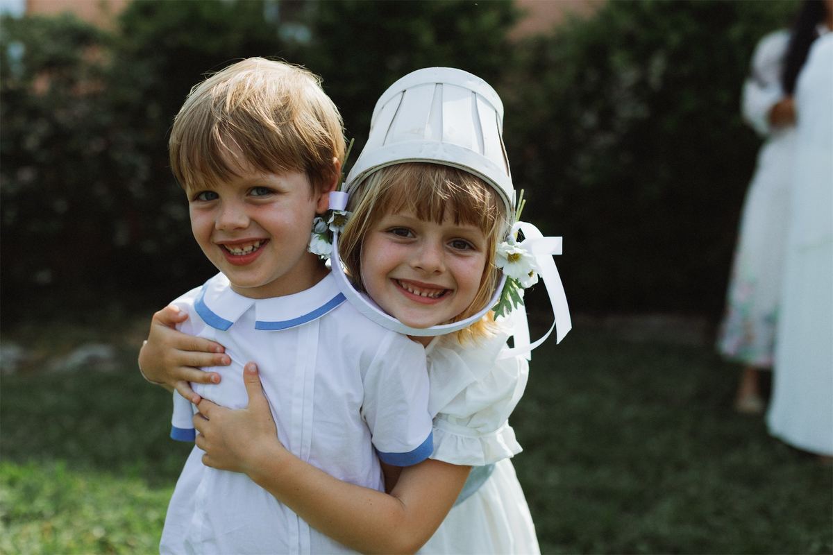 Flower girl in a bonnet hugging the page boy on the grass before the ceremony