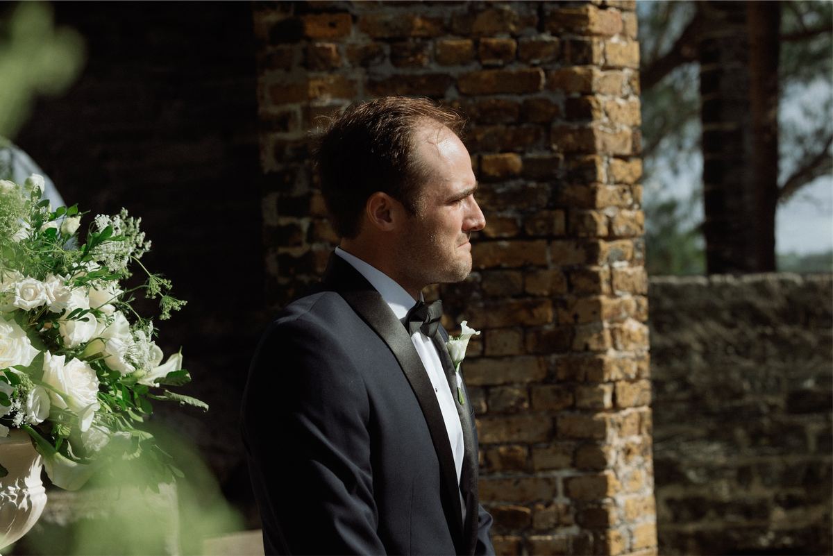 Groom in a navy tuxedo waiting by a brick pillar beside white ceremony flowers