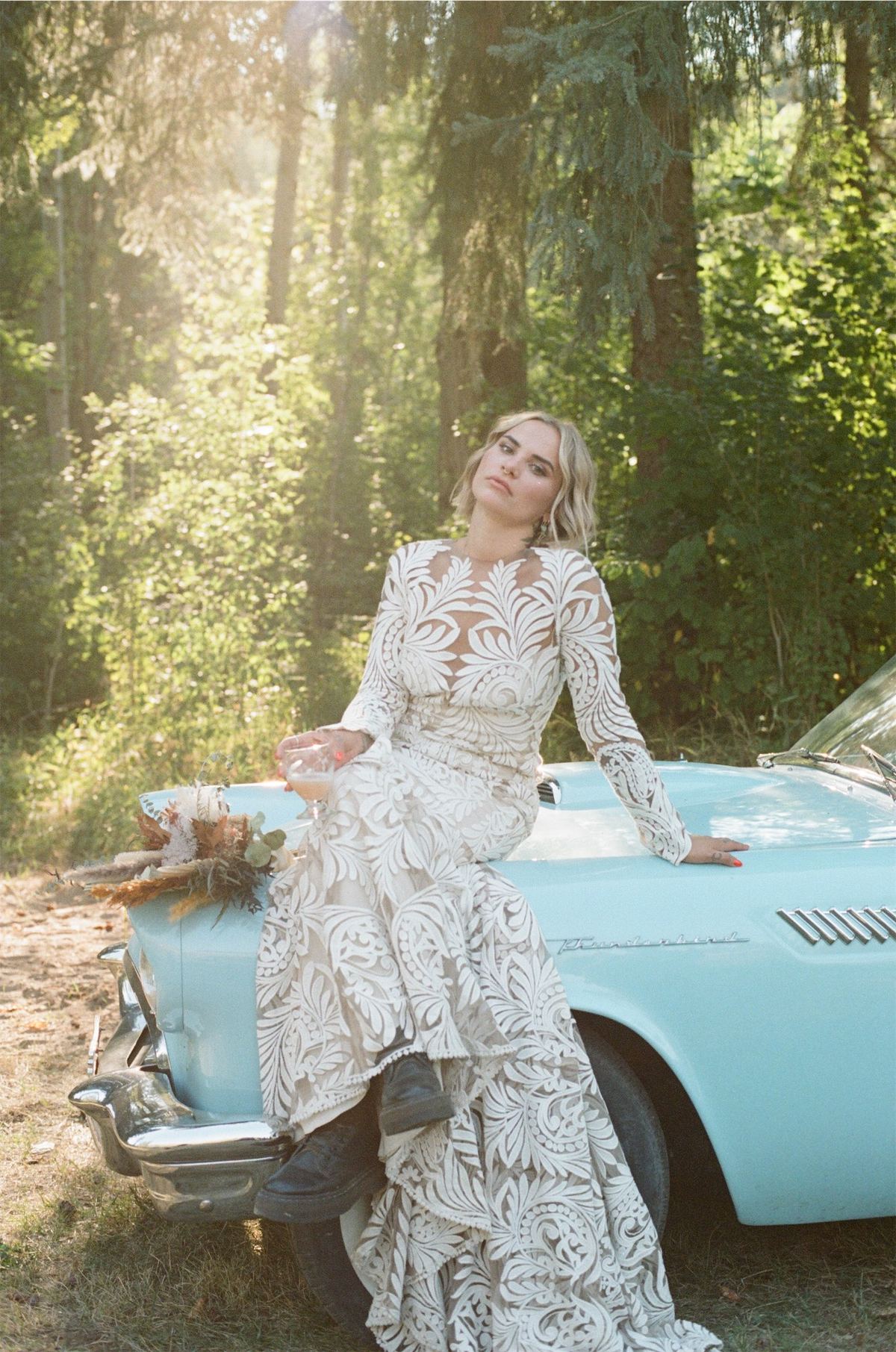 Bride leaning against a turquoise vintage convertible in golden forest light