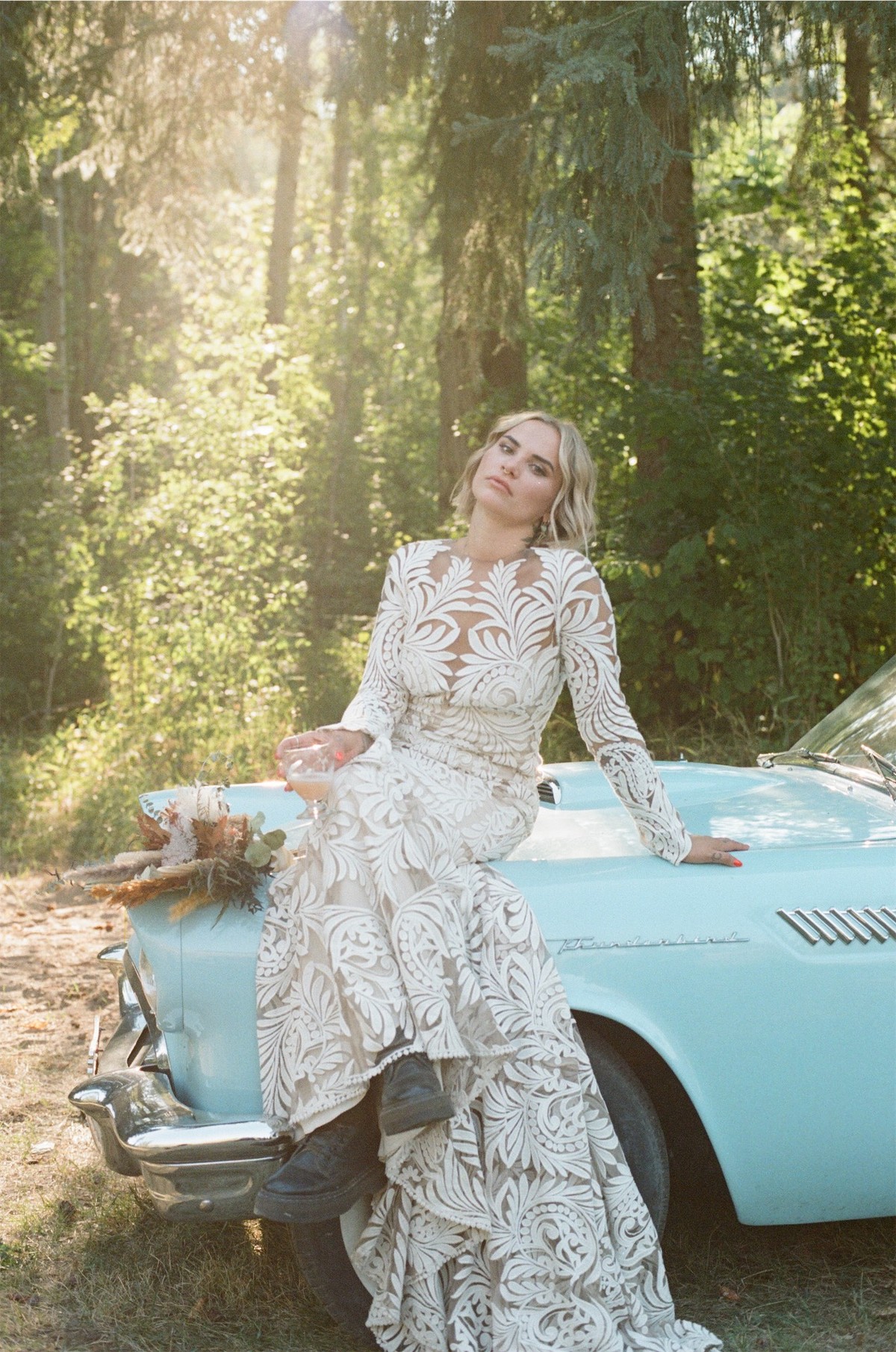 Bride leaning against a turquoise vintage convertible in golden forest light