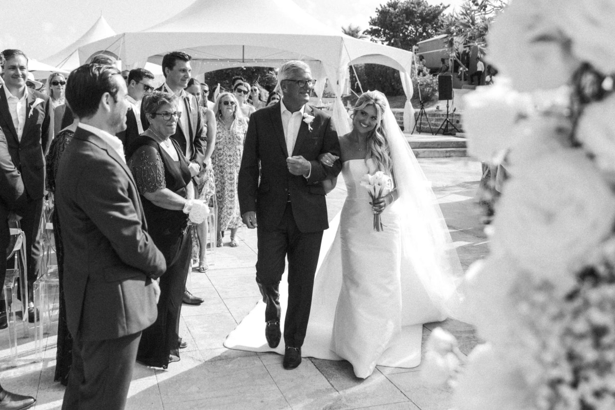 Paige walks down the aisle on her father's arm past the standing guests on Hawkins Island, Bermuda, black and white