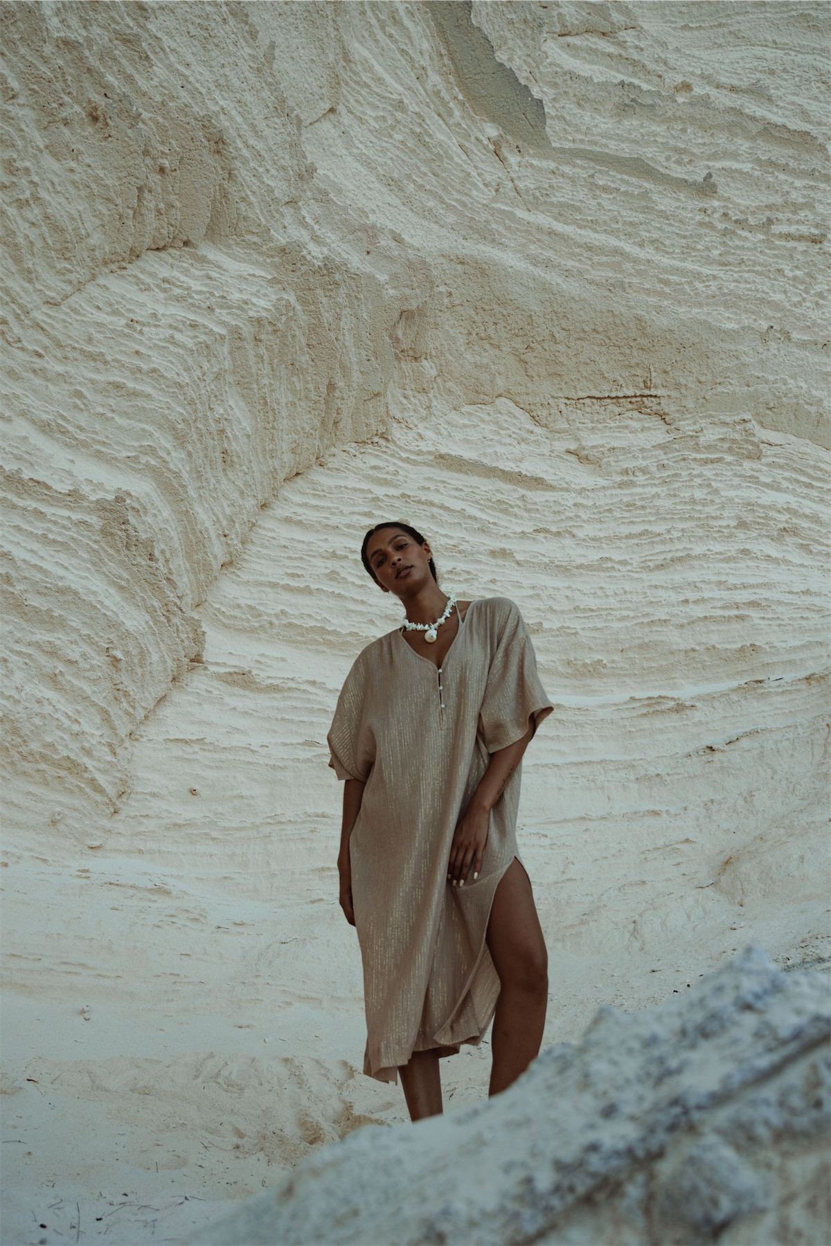 Model in a sand coloured shift dress against a wall of pale rock