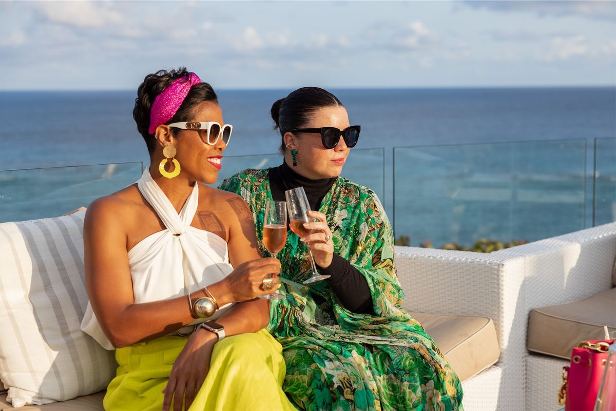 Two women in bold prints toasting with wine on a terrace sofa above the ocean