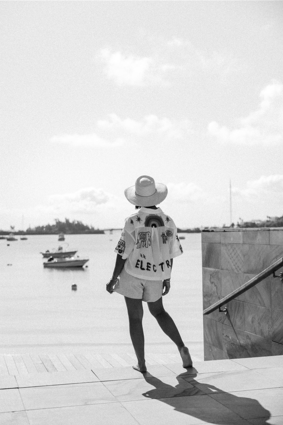 Black and white of a model in a straw hat walking a dock above a boat-filled harbour