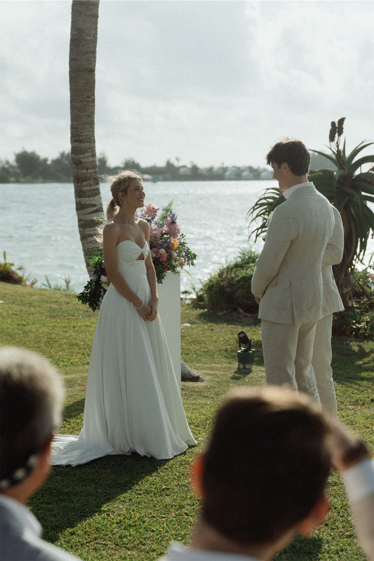Bride and groom exchanging vows on an oceanfront lawn seen between guests