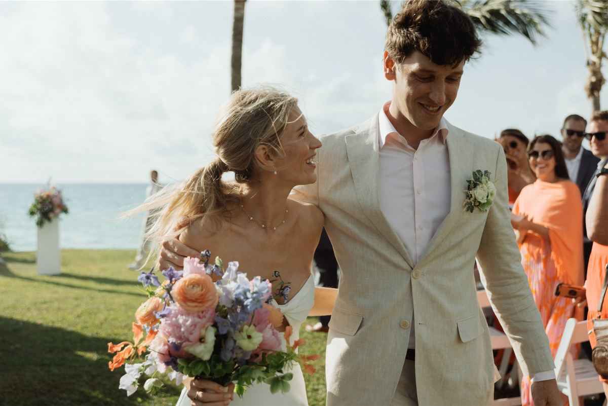 Newlyweds walking arm in arm past cheering guests after their seaside ceremony