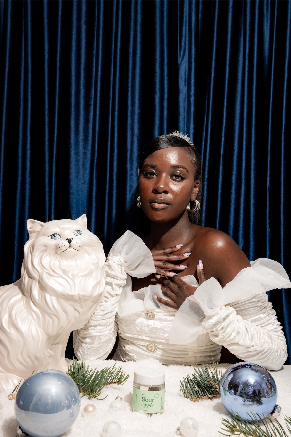 Model seated with a porcelain cat and disco balls before a blue velvet curtain
