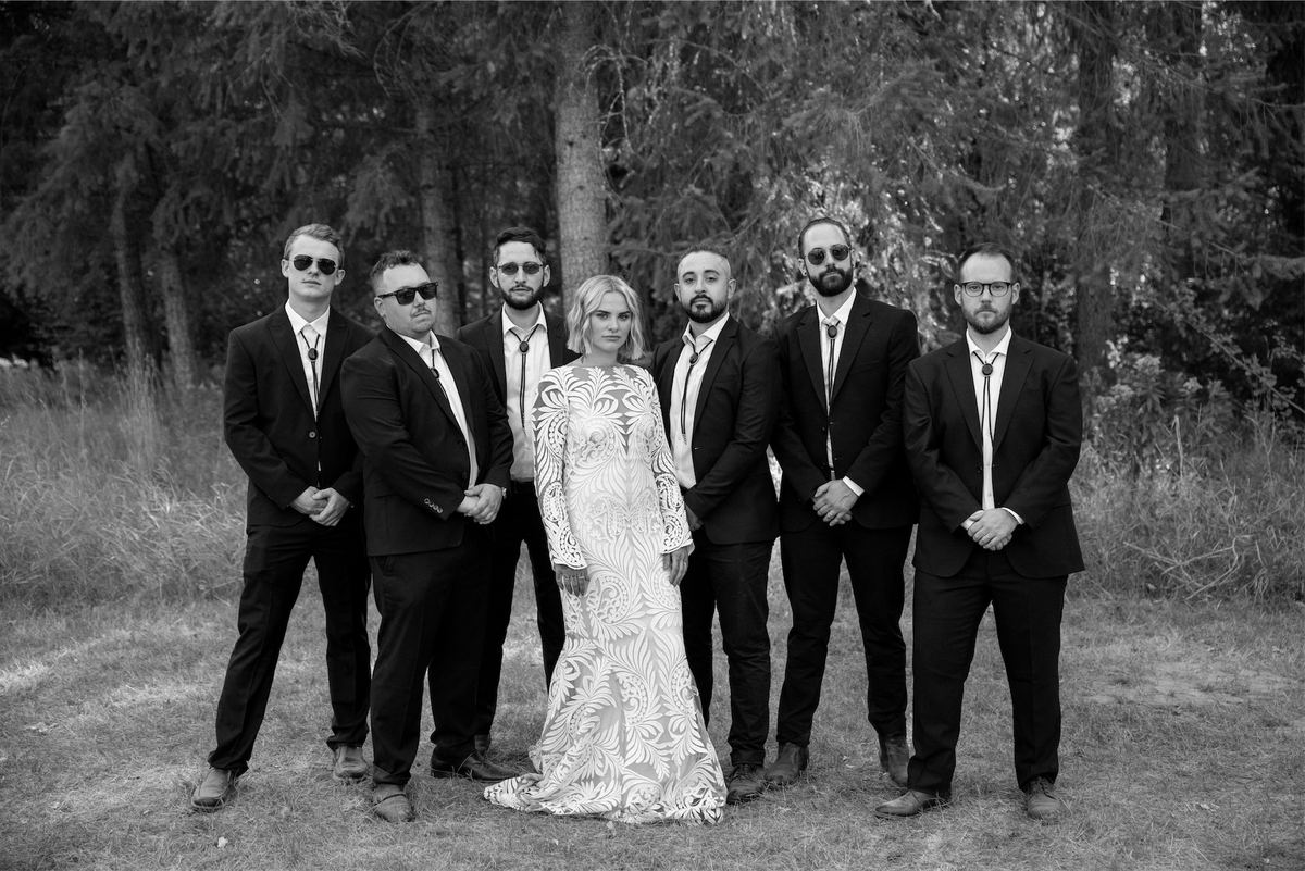Black and white of the bride flanked by groomsmen in black suits among the trees