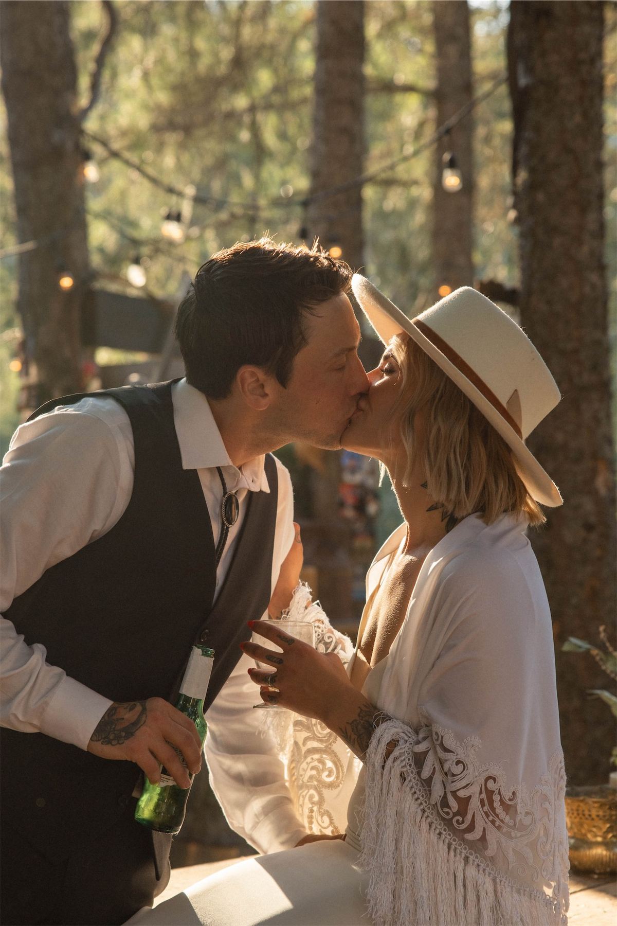 Bride in a wide-brim hat kissing the groom at their woodland reception