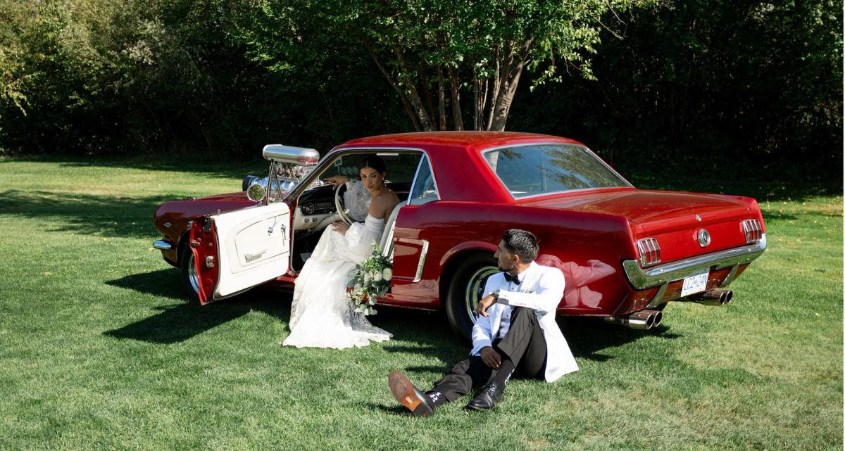 Groom sitting on the grass beside a red vintage Mustang while the bride leans in