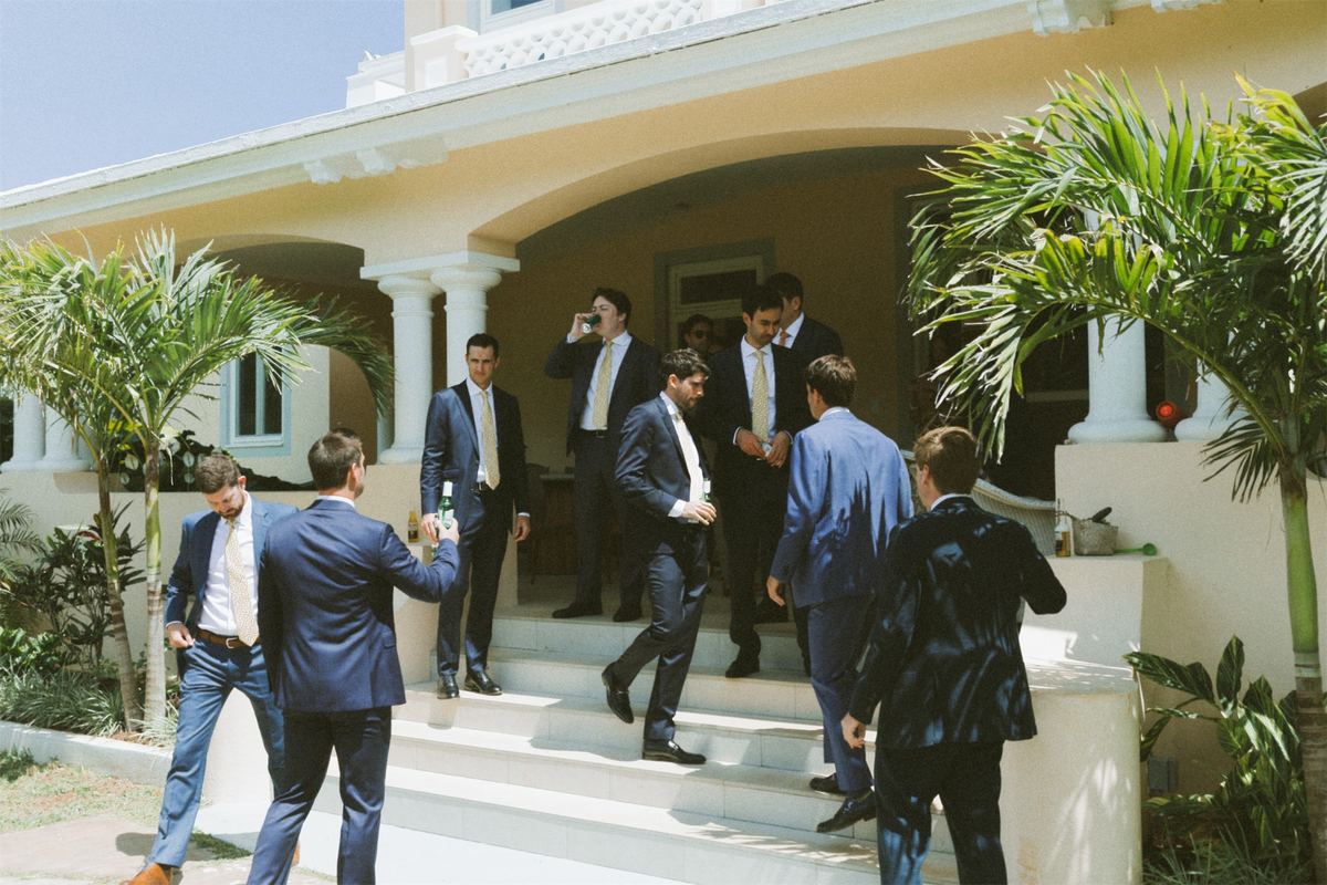 Groomsmen in suits joking with each other on the steps of a pastel villa