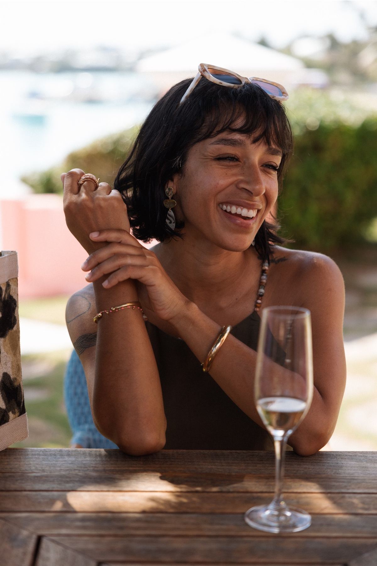 Model laughing over a glass of wine at a sunlit outdoor table, wearing stacked bracelets