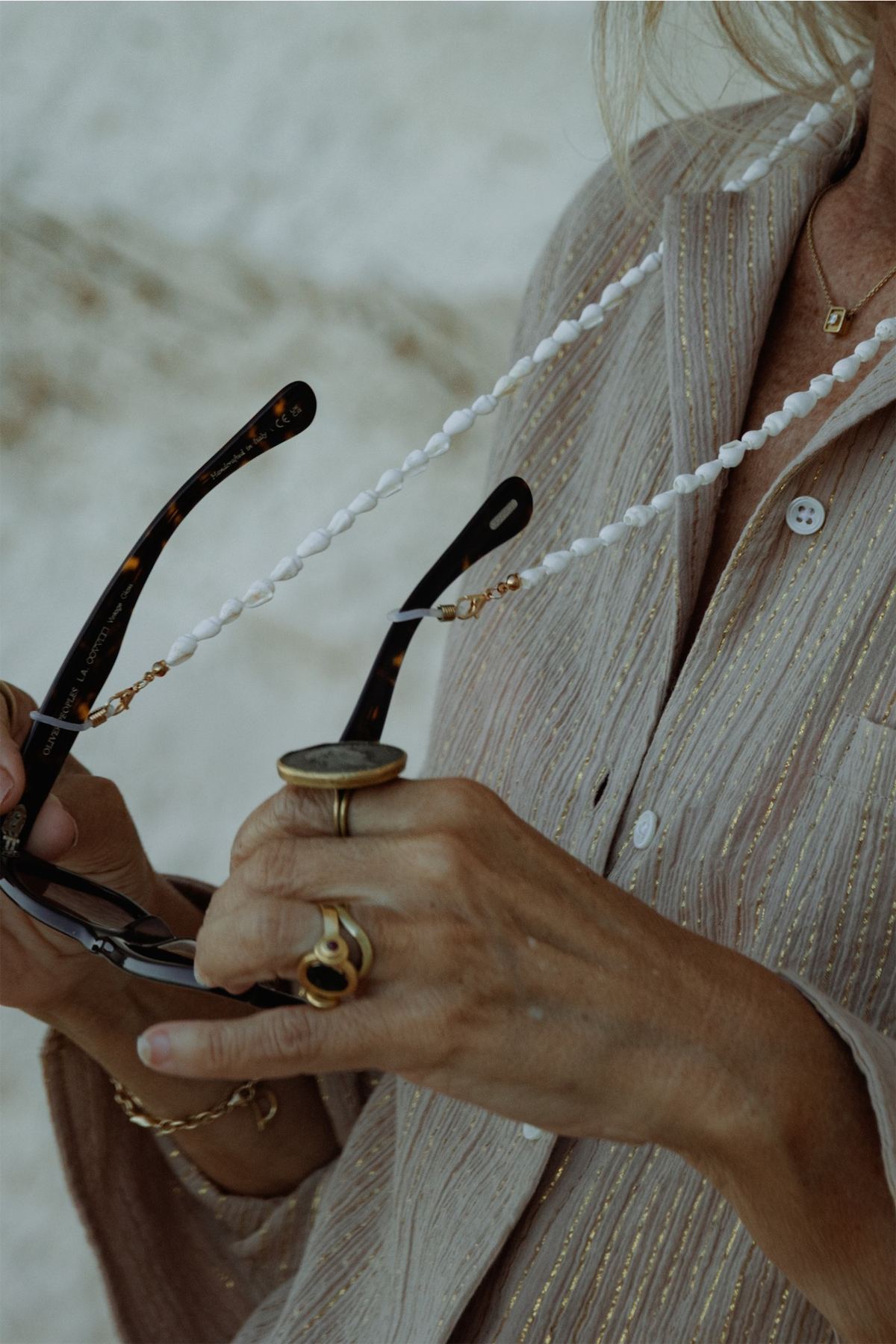 Detail of a ringed hand holding sunglasses on a beaded chain over a linen shirt