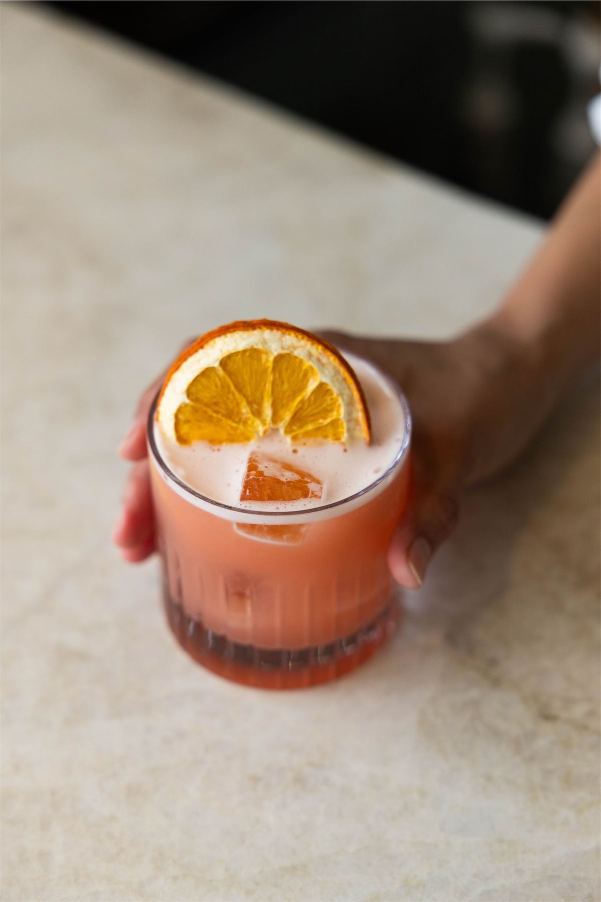Cocktail garnished with an orange wheel held on a pale marble bar top