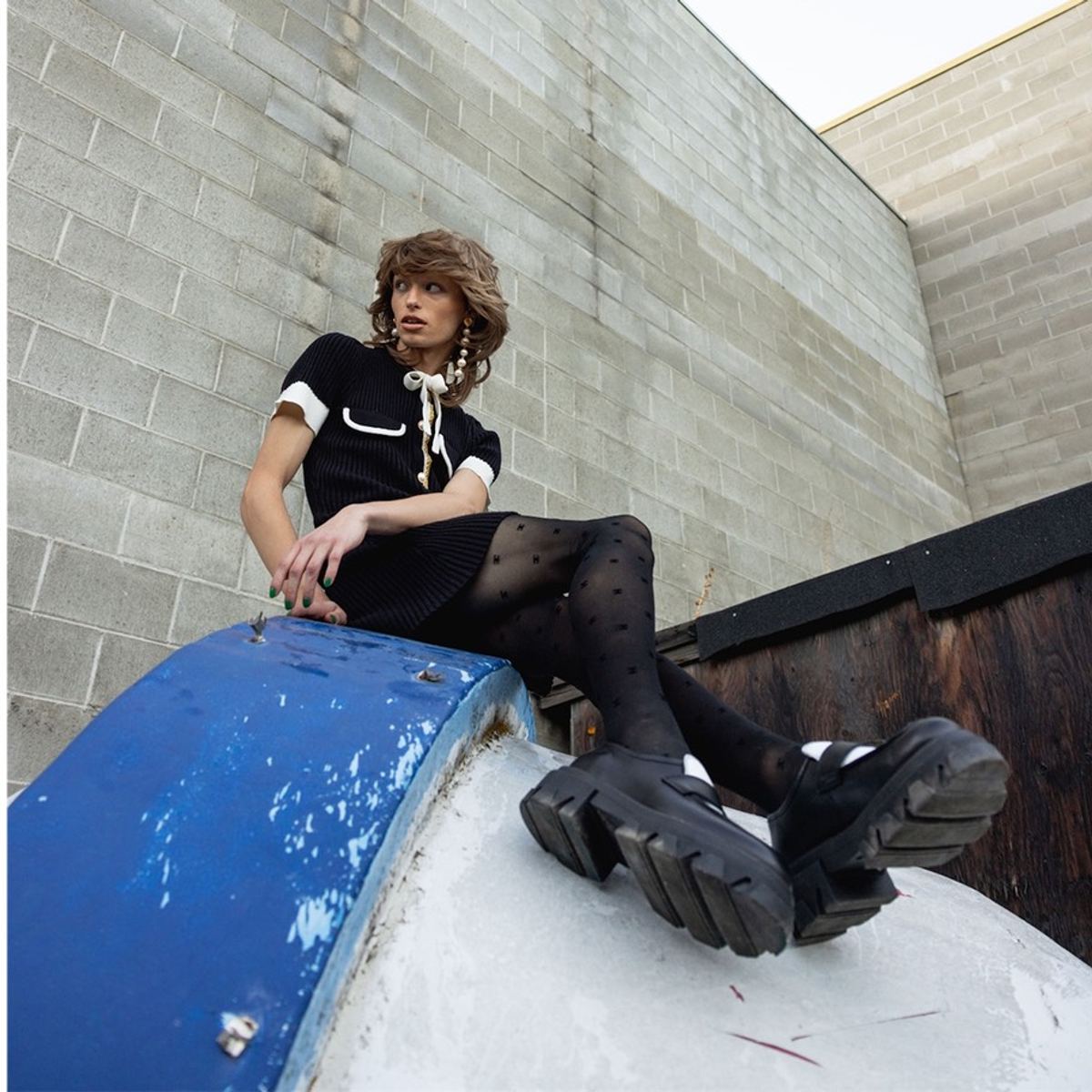 Model in a black dress and platform boots seated on a blue dumpster in an alley