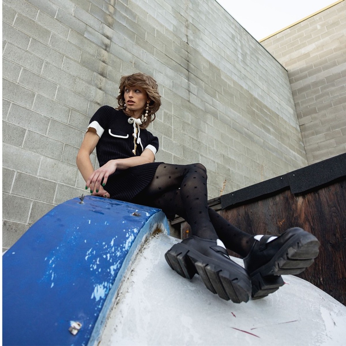 Model in a black dress and platform boots seated on a blue dumpster in an alley