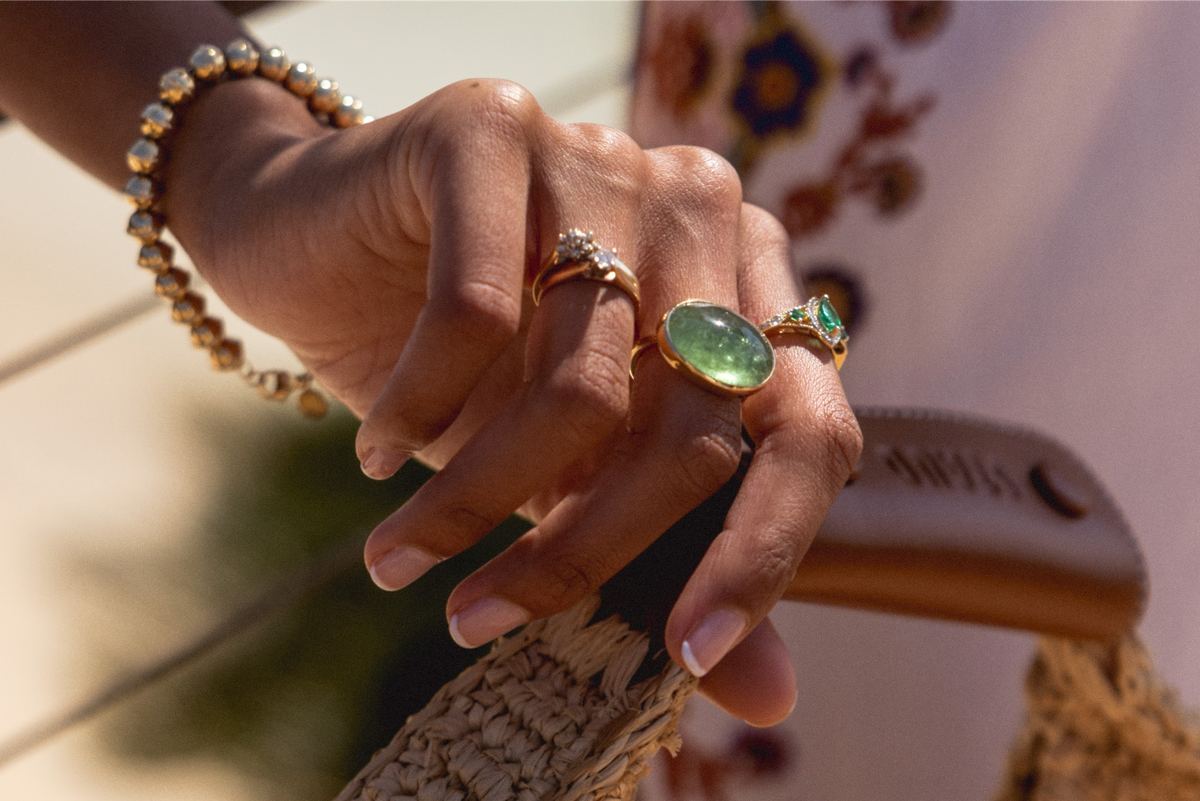 Detail of stacked gold and green gemstone rings on a hand above a straw bag