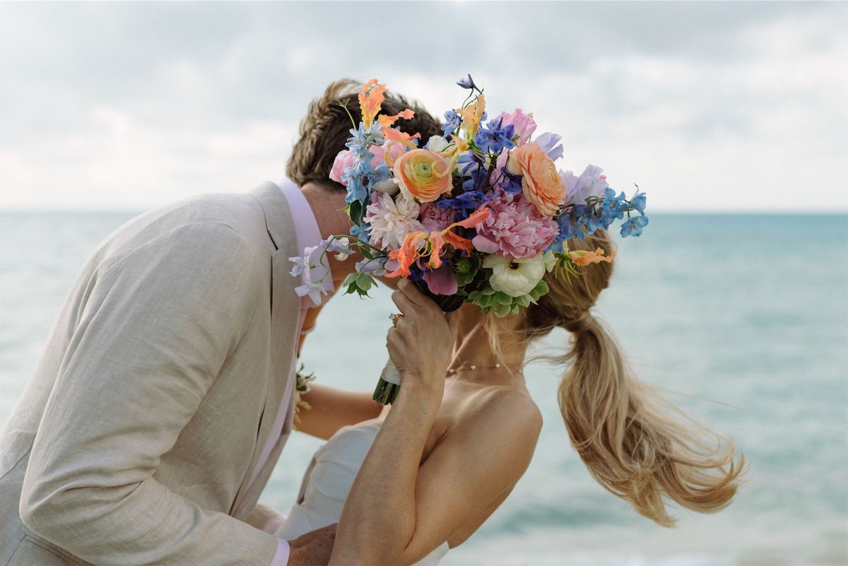 Groom kissing his bride behind her pastel bouquet with the sea behind them