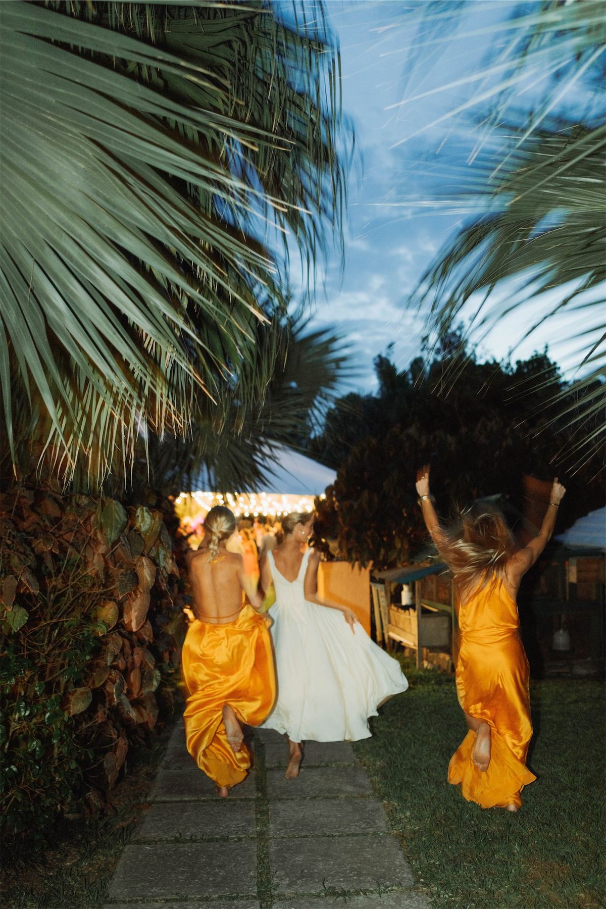 Two bridesmaids in marigold dresses leaping beside the bride under the palms