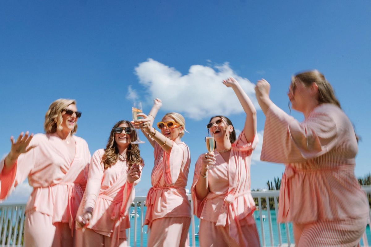 Bridesmaids in pink robes cheering with champagne under a blue Bermuda sky