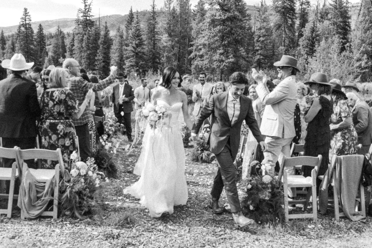 Devin and Owen walk back up the aisle as guests in cowboy hats cheer at a mountain ranch among the aspens, black and white