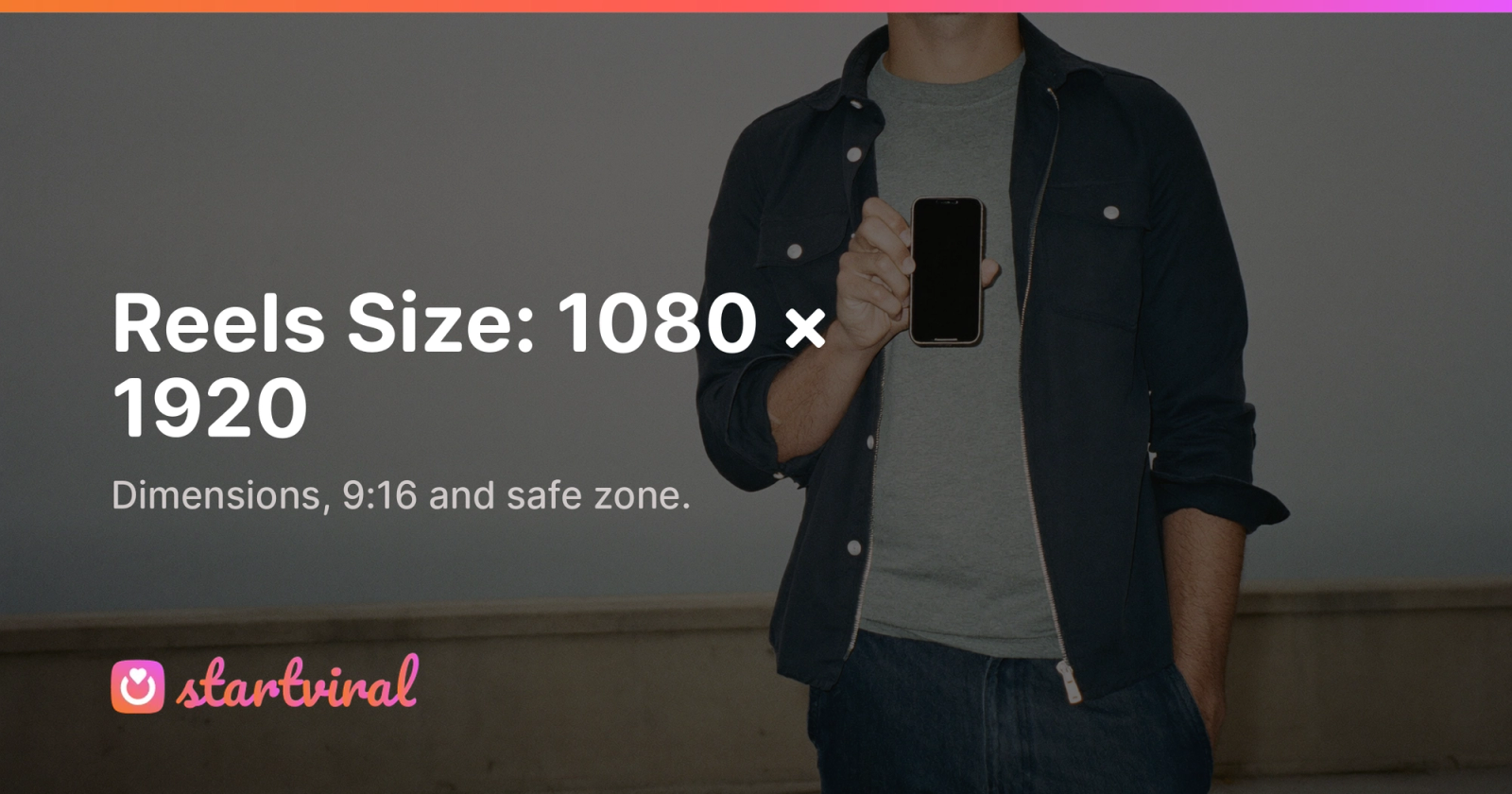 Man in a navy overshirt holding his smartphone upright against a pale wall, as if framing a video.