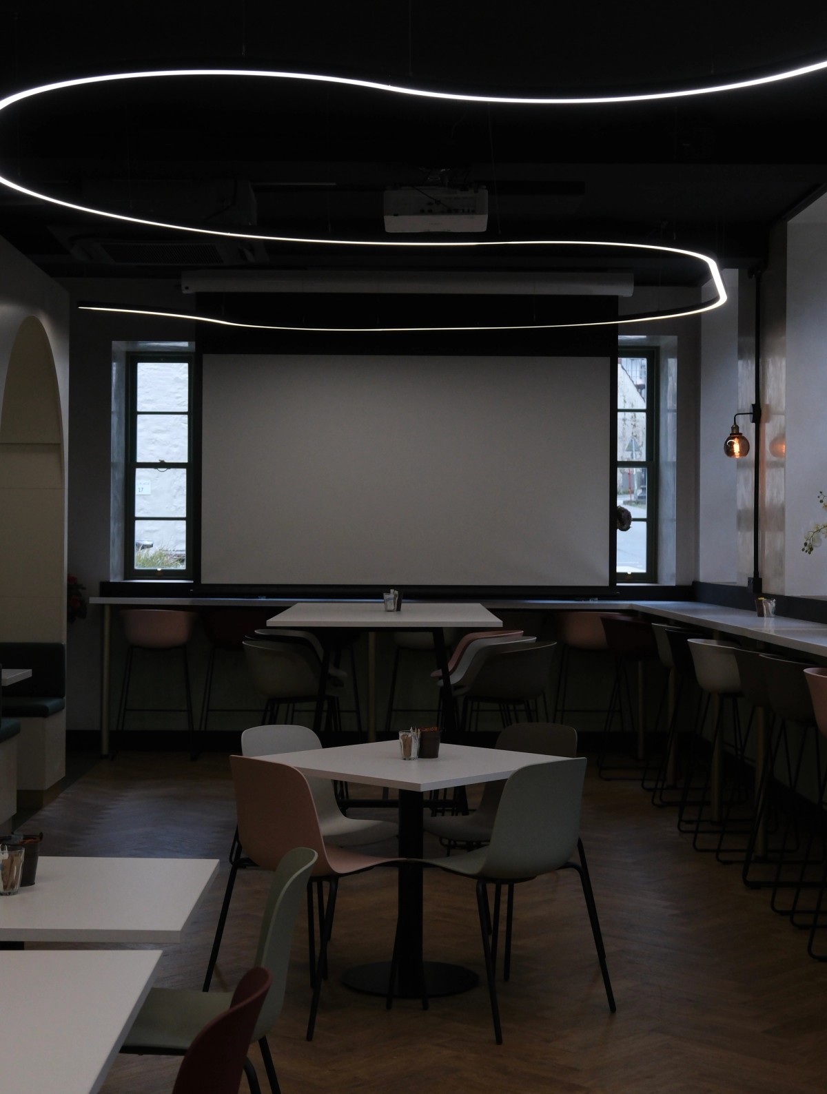 Modern cafe interior with a large projection screen, tables, chairs, and wavy ceiling lights.