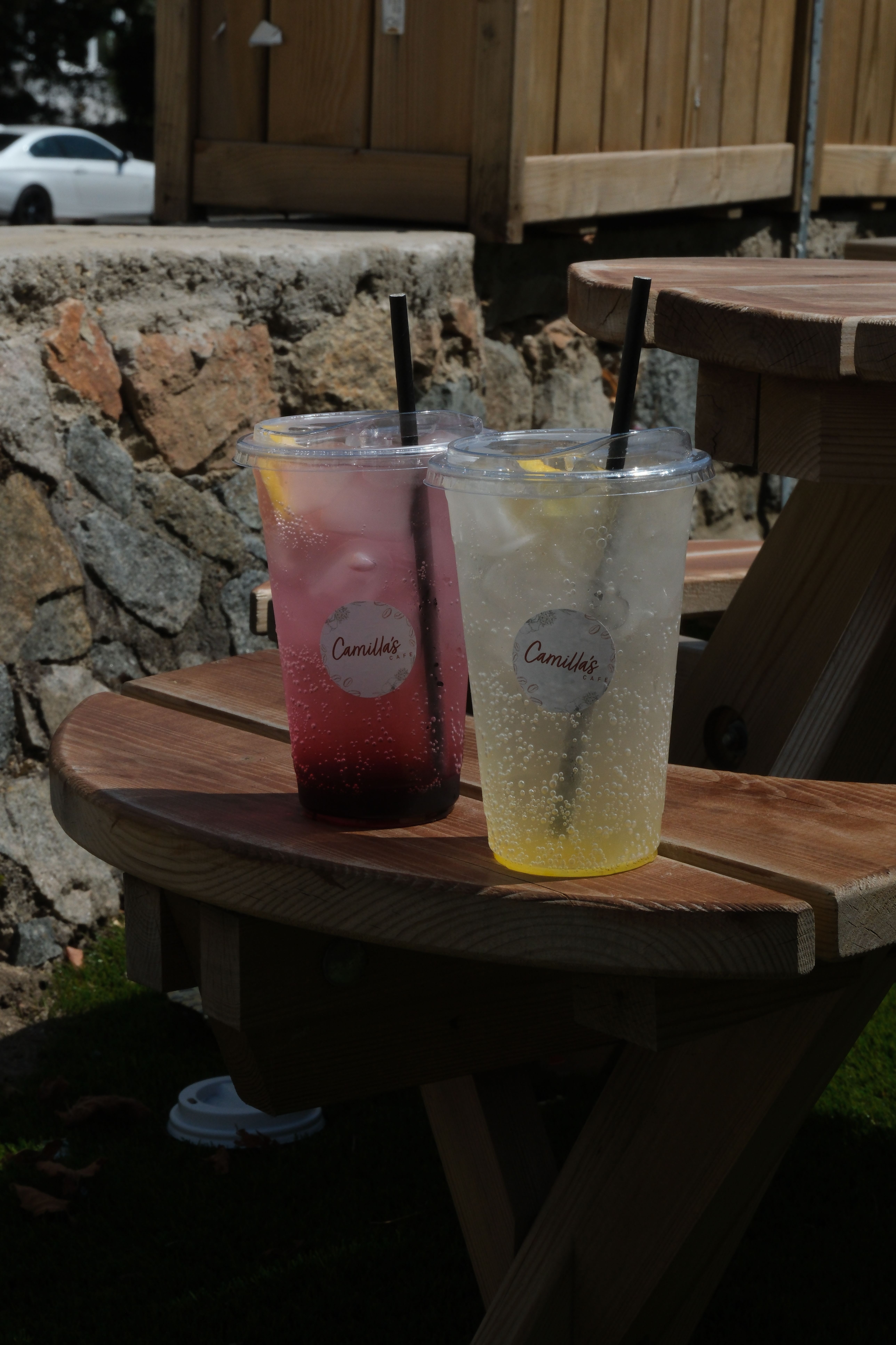Two sparkling lemonades in clear plastic cups, one pink and one yellow with a lemon slice, sitting on a wooden picnic table outdoors.